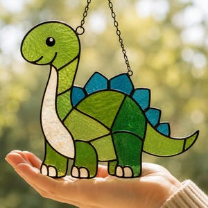 Pode incluir: Um apanhador de sol de vitral em forma de dinossauro com pescoço comprido. O corpo é construído com vários tons de vidro verde, apresentando um peito branco e uma fila de placas triangulares azuis nas costas, unidas por metal preto.