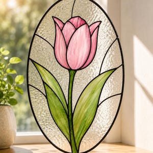 May include: An oval stained glass suncatcher featuring a single pink tulip with green leaves. The flower is composed of pink glass segments with dark lead lines, set against a textured, clear glass background within a black metal frame.