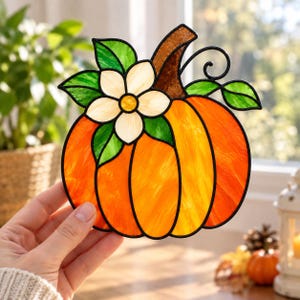 May include: A stained glass pumpkin suncatcher featuring vibrant orange and yellow glass segments with black lead lines. The design includes a brown stem, green leaves, and a white flower with a yellow centre, all framed by a black metal border.