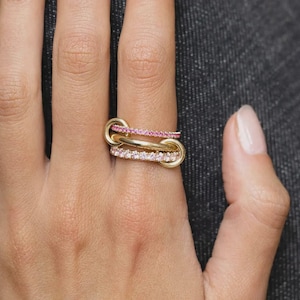 May include: A gold ring featuring three interconnected bands. The top band is set with small pink stones, the middle band is a plain polished gold link, and the bottom band is set with small clear white stones, creating a layered appearance.