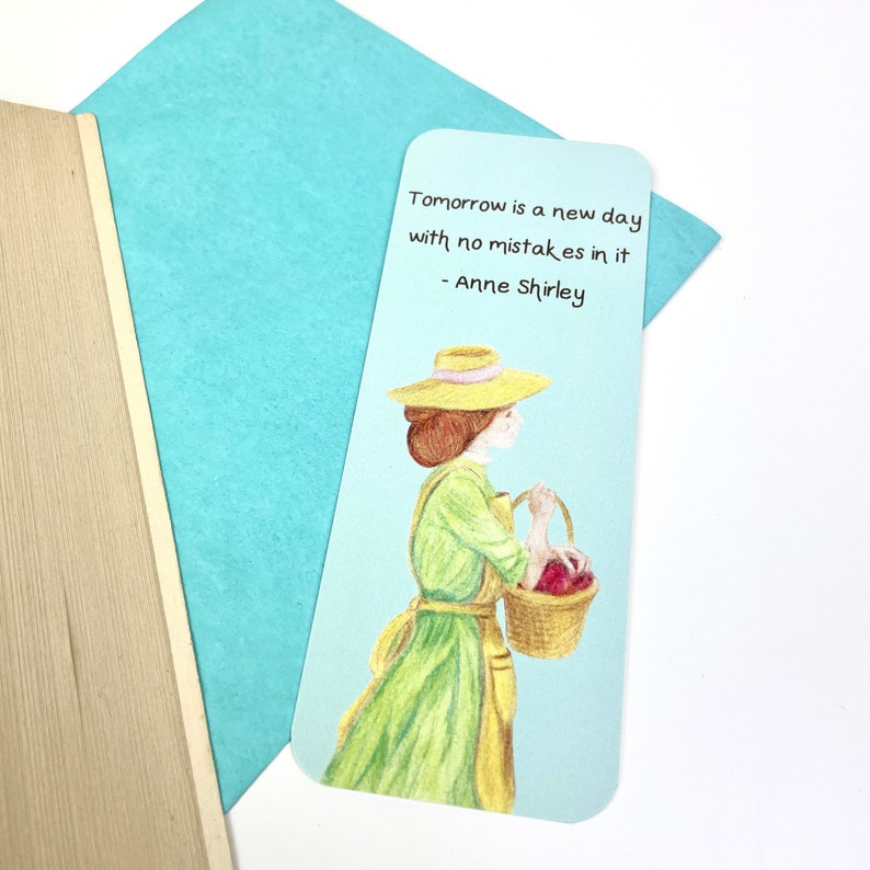 May include: A bookmark featuring a woman in a green dress and straw hat carrying a basket of red apples. The quote "Tomorrow is a new day with no mistakes in it - Anne Shirley" is printed on the bookmark.