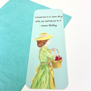May include: A bookmark with a light blue background featuring a woman in a green dress and a straw hat holding a basket of red apples. The quote "Tomorrow is a new day with no mistakes in it - Anne Shirley" is printed on the bookmark.