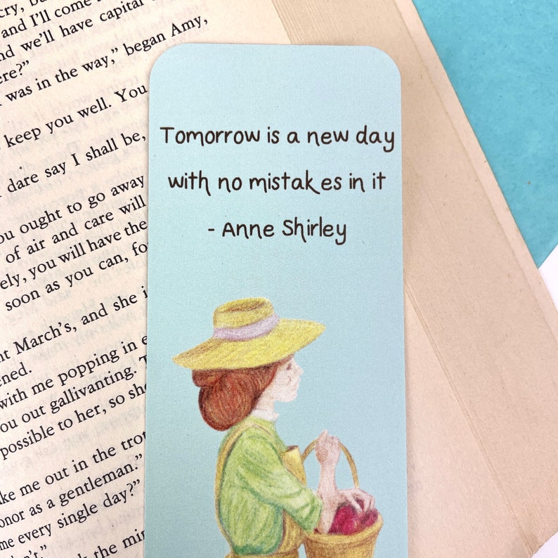 May include: A blue and white bookmark featuring a quote by Anne Shirley: "Tomorrow is a new day with no mistakes in it." The quote is illustrated with a drawing of a woman in a straw hat holding a basket of apples.