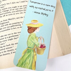 May include: A blue and white bookmark featuring a hand-drawn illustration of a woman in a green dress and a yellow hat carrying a basket of red apples. The quote "Tomorrow is a new day with no mistakes in it - Anne Shirley" is printed on the bookmark.