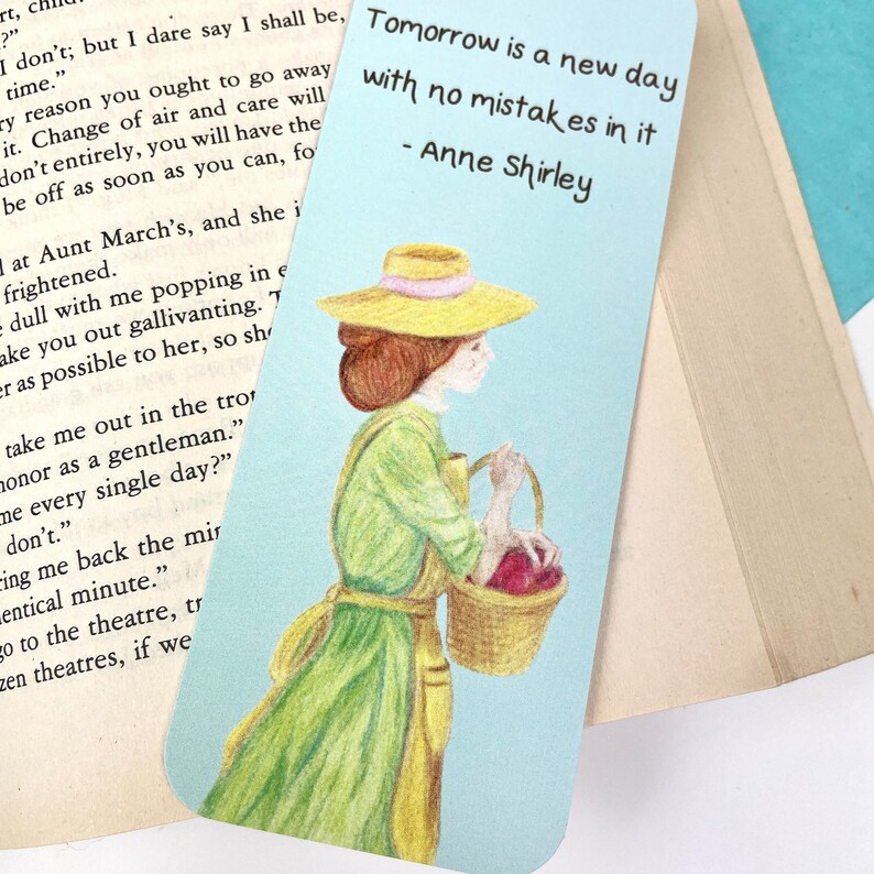 May include: A blue and white bookmark featuring a hand-drawn illustration of a woman in a green dress and a yellow hat carrying a basket of red apples. The quote "Tomorrow is a new day with no mistakes in it - Anne Shirley" is printed on the bookmark.