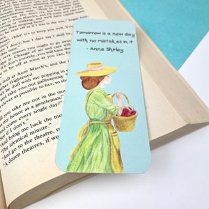 May include: A blue and white bookmark featuring a watercolour illustration of a woman in a green dress and yellow apron carrying a basket of red apples. The text on the bookmark reads "Tomorrow is a new day with no mistakes in it - Anne Shirley".