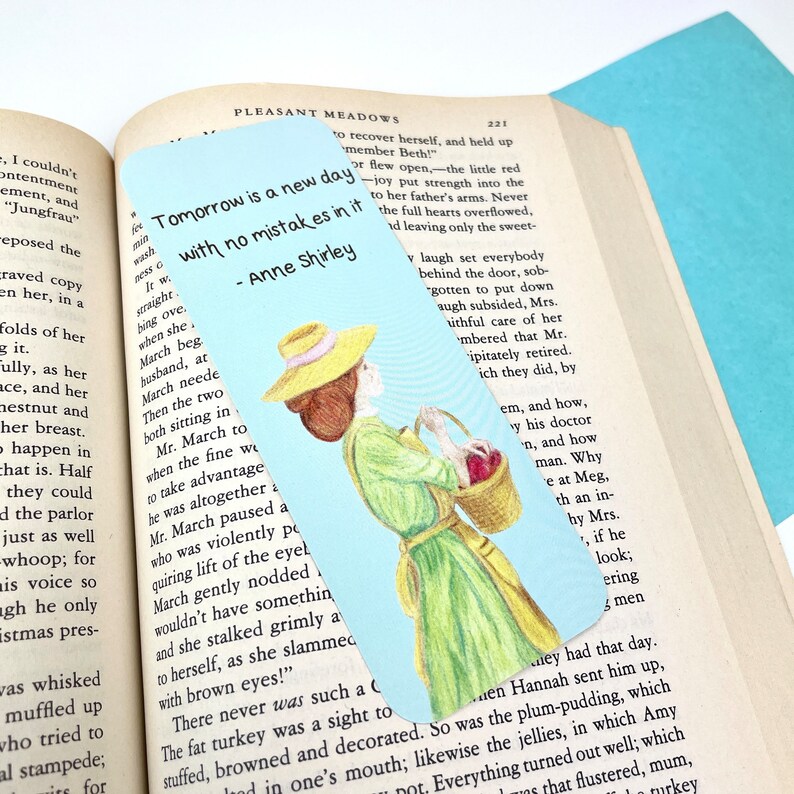May include: A blue bookmark with a hand-drawn illustration of a woman in a yellow hat and green dress carrying a basket of apples. The bookmark features the quote "Tomorrow is a new day with no mistakes in it" - Anne Shirley.