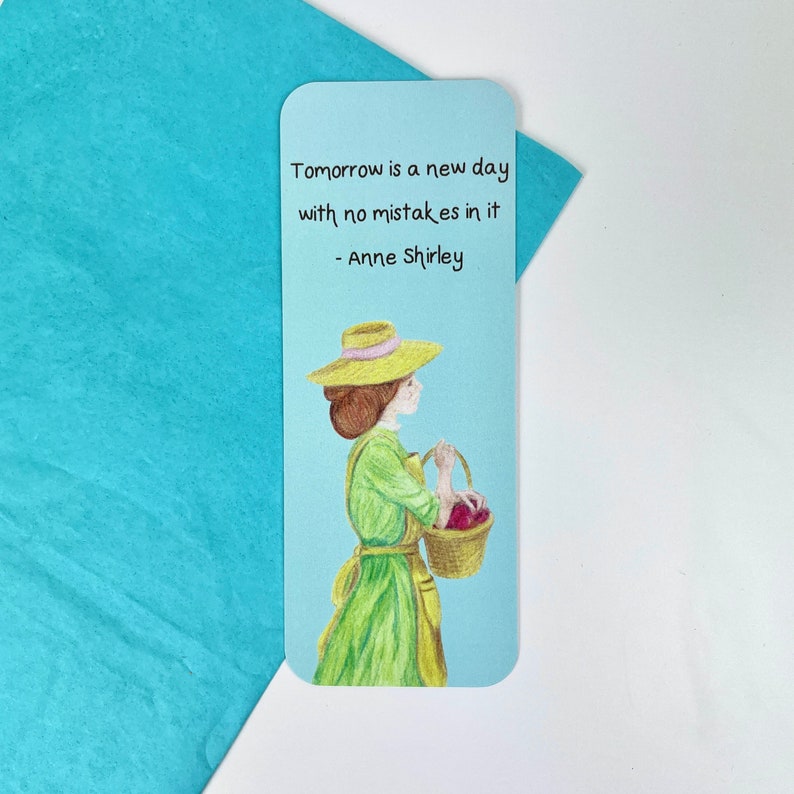 May include: A blue bookmark featuring a quote from Anne Shirley: "Tomorrow is a new day with no mistakes in it." The quote is illustrated with a woman in a green dress and a straw hat carrying a basket of red apples.