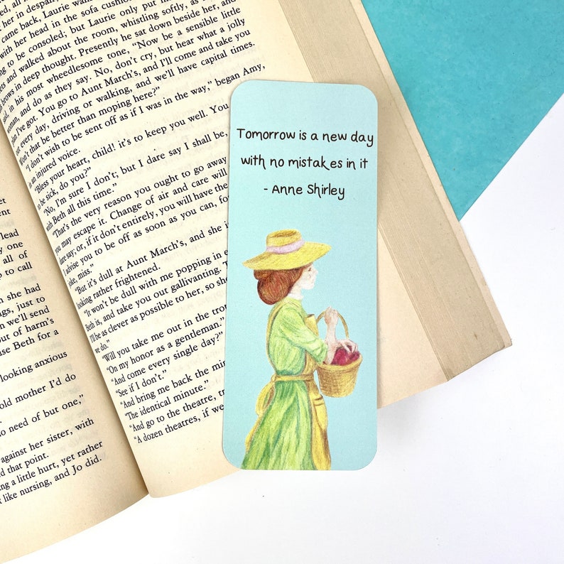 May include: A blue and white bookmark featuring a quote by Anne Shirley: "Tomorrow is a new day with no mistakes in it". The bookmark depicts a watercolour illustration of a woman in a green dress and a straw hat, carrying a basket of red apples.