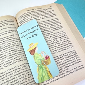 May include: A blue and white bookmark featuring a quote from Anne Shirley, "Tomorrow is a new day with no mistakes in it." The quote is printed in black text. The bookmark features a hand-drawn illustration of a woman in a yellow hat and green dress holding a basket of red apples.