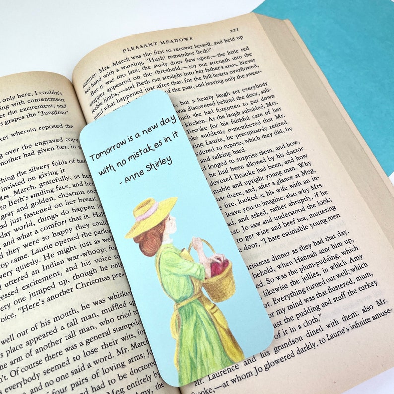 May include: A blue and white bookmark featuring a quote from Anne Shirley, "Tomorrow is a new day with no mistakes in it." The quote is printed in black text. The bookmark features a hand-drawn illustration of a woman in a yellow hat and green dress holding a basket of red apples.
