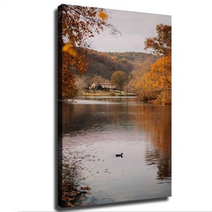 May include: A vertical canvas wall art print depicting a serene lake scene during autumn. A lone duck swims in the calm water, reflecting the surrounding trees with vibrant orange and brown foliage. A house sits on the distant shore near hills.