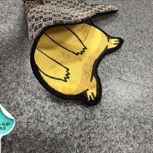 May include: A yellow rug with a black outline and a graphic of a creature, featuring a black and white checkered non-slip sole. The rug is partially folded to reveal the underside, which includes a circular label with the text NON-SLIP SOLE.