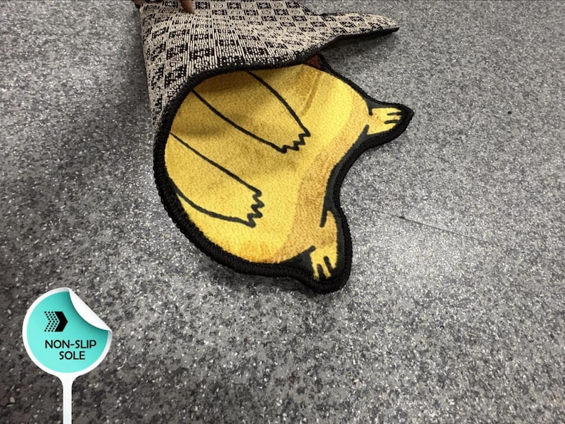 May include: A yellow rug with a black outline and a graphic of a creature, featuring a black and white checkered non-slip sole. The rug is partially folded to reveal the underside, which includes a circular label with the text NON-SLIP SOLE.