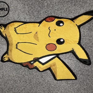 May include: A yellow, black, and red rug shaped like the character Pikachu. The rug features the character's iconic pointed ears with black tips, round red cheeks, and a small mouth. A circular black stamp with the word SAMPLE is in the top left.