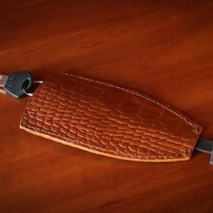 May include: A brown leather key fob with a textured crocodile skin pattern. The key fob is shaped like a shield and features a metal key ring attached to a silver-coloured metal key on one end and a small black leather pull tab on the other end.