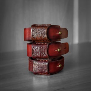 Op de afbeelding: Een stapel van drie donkerrode leren armbanden, elk met een centraal in reliëf gemaakt cirkelvormig ontwerp. De bovenste en onderste armbanden tonen een boommotief, terwijl de middelste een geometrisch knooppatroon heeft. Sluiting met een messing knop.