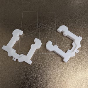 Peut inclure: Deux composants en plastique blanc imprimés en 3D avec un cadre en forme de U, présentant des découpes géométriques complexes, des bords crantés et de petites broches saillantes sur le dessous. Ces pièces identiques ont une finition mate.