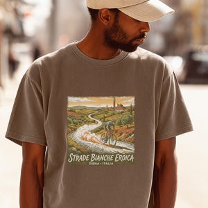 May include: A taupe short-sleeve t-shirt featuring a graphic illustration of a cyclist riding on a gravel road through a landscape with buildings in the distance. The text reads Strade Bianche Eroica SIENA ITALIA in a white serif font.