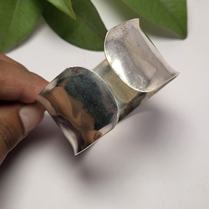 May include: A wide, polished silver-tone cuff bracelet with a smooth, reflective surface and a curved, open-ended design. The metal band features a subtle wave-like contour along its edges, creating a continuous, flowing shape that wraps around.