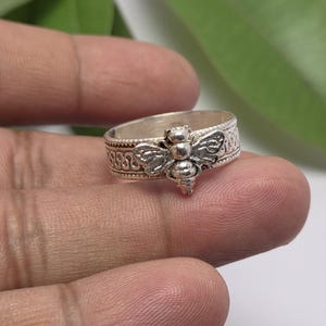 May include: A silver-toned ring featuring a prominent bee motif centered on the band. The band displays an intricate, embossed pattern that wraps around the circumference, providing a textured appearance to the metallic surface of the jewelry.