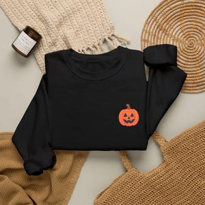 Könnte beinhalten: Ein schwarzes Langarm-Sweatshirt mit einem kleinen, gestickten orangefarbenen Jack-o'-Lantern-Kürbis auf der linken Brust. Das Kleidungsstück hat einen Rundhalsausschnitt, lange Ärmel mit gerippten Bündchen und ein schlichtes Design.