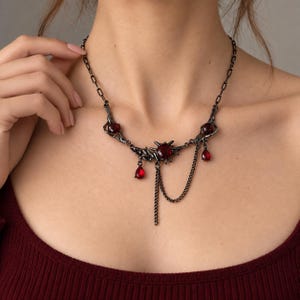 May include: A dark gunmetal chain necklace featuring a central star-shaped setting with a deep red gemstone, accented by two additional red teardrop-shaped pendants and a secondary delicate chain drape hanging from the main centrepiece.