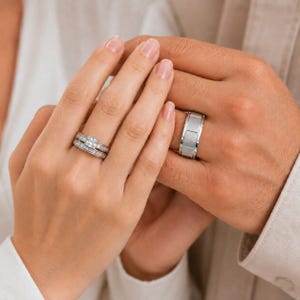 Könnte beinhalten: Ein silberfarbenes Verlobungsring-Set mit einem zentralen quadratischen Stein, der von Baguette-geschliffenen Steinen am Ringband umgeben ist, sowie einem breiten, gebürsteten silberfarbenen Ehering mit segmentiertem, rechteckigem Muster.