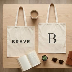 May include: Two off-white canvas tote bags with black lettering. One bag features the word BRAVE in a sans-serif font, while the other displays a large serif letter B. The bags have two shoulder straps each and are made of a plain fabric.