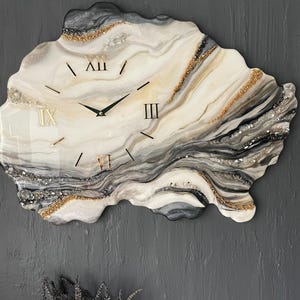 Può includere: Un orologio da parete di forma irregolare ispirato ai geodi, caratterizzato da un motivo marmorizzato con vortici nei toni del crema, bianco, grigio e nero. Il quadrante presenta numeri romani dorati alle ore tre, sei, nove e dodici, con lancette nere.