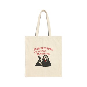 May include: A beige canvas tote bag featuring a graphic of a black-robed figure with a white mask holding a knife, lying down. Above the figure, red text reads: PEER PRESSURE, I'M FAR TOO SENSITIVE! The bag has two long shoulder straps for carrying.