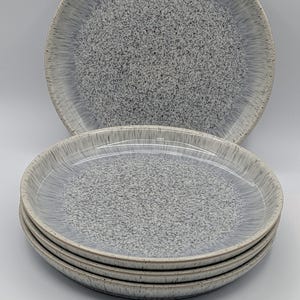 Peut inclure: Une pile de quatre assiettes rondes en céramique présentant un motif moucheté gris et blanc. Le centre de chaque assiette présente un motif dense de taches gris foncé qui s'estompe en un dégradé gris clair et blanc vers le bord surélevé.