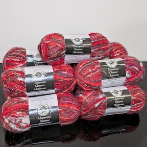 May include: A collection of six skeins of Loops & Threads Glamorous yarn in a variegated red, black, and silver metallic colorway. Each skein features a white label with the text Glamorous, Glamour, Glamoroso, and 53 yds / 49 m printed clearly.
