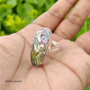 May include: An oval-shaped ring featuring a polished abalone shell inlay with iridescent green, pink, and blue hues. The shell is set within a smooth, silver-toned metal band that features a split shank design on the side of the ring.