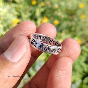 May include: A silver band ring featuring embossed black Tibetan script characters around the entire outer surface. The metal has a polished finish with darkened recessed areas that highlight the intricate lettering and symbols engraved on the band.
