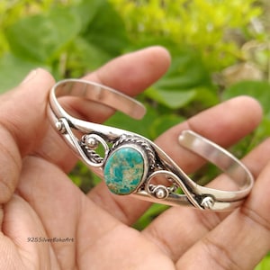May include: A sterling silver cuff bracelet featuring a central oval turquoise stone with natural veining. The metal band displays a polished finish with decorative twisted wire detailing and small silver spheres flanking the stone setting.
