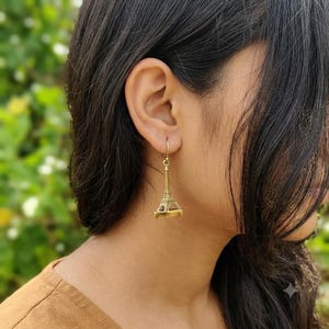 May include: A gold-toned metal dangle earring shaped like the Eiffel Tower. The charm features a detailed, three-dimensional architectural design with a pointed spire and arched base, suspended from a simple metal hook wire that passes through the ear.
