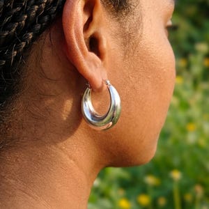 May include: A thick, polished silver-tone hoop earring with a smooth, rounded surface. The earring features a classic circular design with a hinged closure mechanism, reflecting light across its metallic, curved exterior surface.