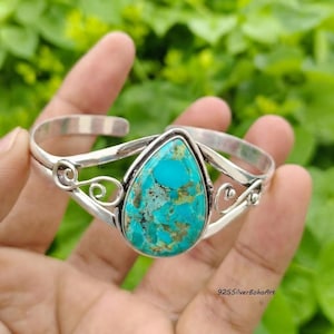 May include: A silver cuff bracelet featuring a large, pear-shaped turquoise stone with brown matrix veining. The metal band displays openwork scroll designs on both sides of the central gemstone, creating a decorative and detailed metal frame.