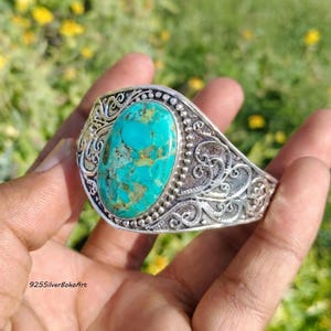 May include: A silver cuff bracelet featuring a large, oval-shaped turquoise stone with brown matrix veining at the centre. The metal band is decorated with intricate, raised scrollwork and bead detailing surrounding the central gemstone.