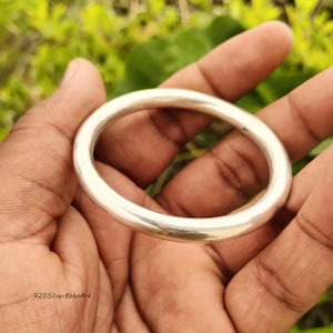 Peut inclure: Un bracelet jonc en argent poli et lisse avec un profil circulaire arrondi. La surface métallique est réfléchissante et d'une épaisseur uniforme, formant une boucle simple et continue. Le texte 925SilverBohoArt est visible dans le coin.