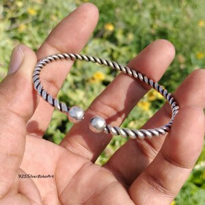 Può includere: Un bracciale a fascia in metallo color argento caratterizzato da una texture intrecciata simile a una corda lungo tutta la banda. Il bracciale ha un design aperto, rifinito con una perla sferica liscia e lucida su ciascuna estremità.