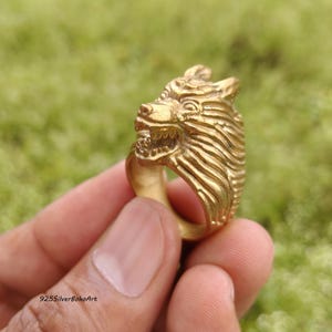 Könnte beinhalten: Ein goldfarbener Metallring mit einem detaillierten, dreidimensionalen Wolfskopf-Design. Der Wolf ist mit leicht geöffnetem Maul dargestellt, das Zähne zeigt, und weist eine texturierte Fellzeichnung an den Seiten des Rings auf. Der Text 925SilverBohoArt ist sichtbar.
