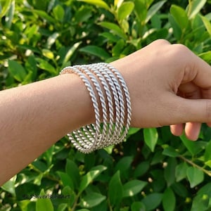 Può includere: Un set di cinque bracciali rigidi color argento, ognuno caratterizzato da una texture intrecciata simile a una corda. I bracciali sono indossati insieme, creando un aspetto uniforme con una finitura metallica che riflette la luce sulla superficie.