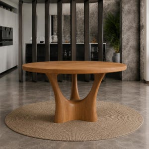 Può includere: Un tavolo da pranzo rotondo in legno con una finitura liscia color marrone chiaro e venature del legno visibili. Il tavolo presenta una base centrale scultorea con tre gambe curve interconnesse che sostengono il piano circolare.