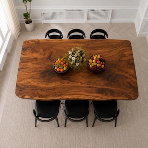 May include: A large rectangular dining table with a natural wood grain finish and live edge sides, surrounded by six black curved chairs. Two bowls filled with assorted fresh fruit and a central floral arrangement sit on the tabletop surface.