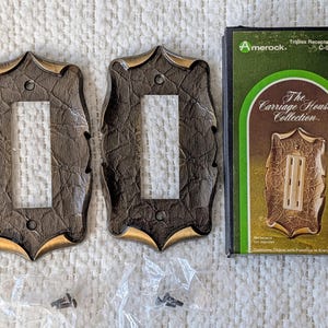 May include: Two brown and gold decorative outlet covers with a textured finish, next to a box labeled "The Carriage House Collection." The box features a green and gold design and the Amerock logo. Includes screws.