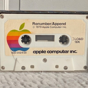 May include: A vintage Apple Computer Inc. cassette tape with the text "Renumber/Append" and the Apple logo. The tape is white with a colorful rainbow apple logo and the text "LOAD 16K".