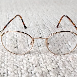 May include: Gold-rimmed eyeglasses with oval lenses. The frames have a thin, delicate design. The temples are patterned with brown and black tortoiseshell. The glasses are set against a textured, off-white background.