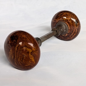 May include: A pair of round door knobs featuring a glossy, marbled brown and tan finish. The two knobs are connected by a central metal spindle, with visible metal collars and small screws securing the assembly together in a functional unit.